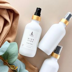 three white bottles with gold caps next to some flowers and a brown cloth on the ground