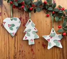 two ceramic christmas tree ornaments hanging from a branch on a wooden table with berries and holly