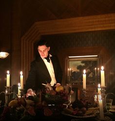 a man in a tuxedo standing at a table with candles and fruit on it