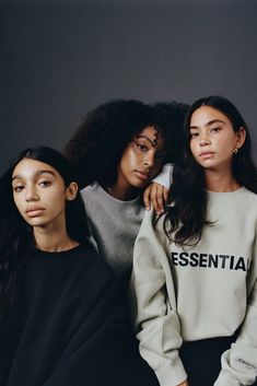 three young women sitting next to each other in front of a gray wall wearing sweatshirts