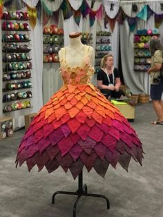 a dress made out of colored paper on a stand in a room with people looking at it