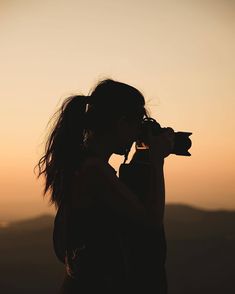 a woman holding a camera up to her face in front of the sun setting behind her