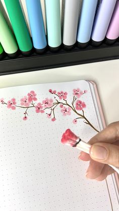 a hand holding a brush near a notebook with pink flowers on it and colored markers in the background