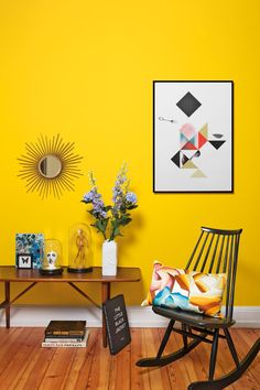 a yellow room with a rocking chair, table and flowers in vases on the floor