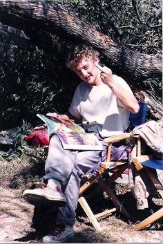 a man sitting in a folding chair reading a magazine under a fallen tree, while holding his hand up to his ear