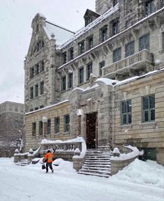 a man walking down a snow covered street in front of a large stone building with many windows