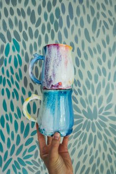 a hand holding a painted mug in front of a wall