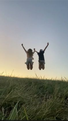 two people jumping in the air on top of a grass covered hill with their arms up