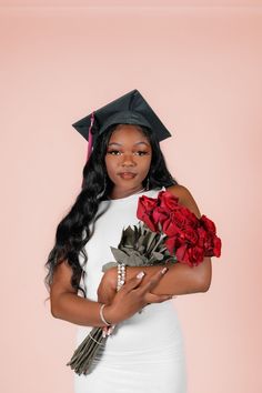 a woman wearing a graduation cap and gown holding flowers in her hands while standing against a pink background