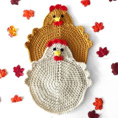 two crocheted roosters sitting next to each other on a white surface surrounded by fall leaves