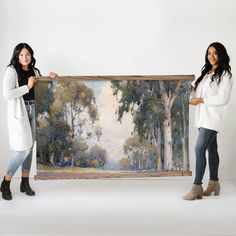 two women standing next to each other holding up a large painting with trees on it