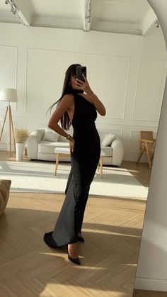 a woman taking a selfie in a room with white walls and wooden floors, wearing a black dress