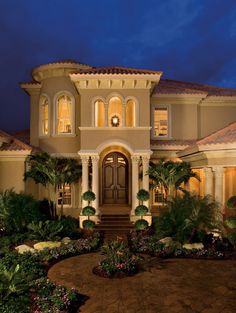 a large house lit up at night with palm trees and shrubs in front of it