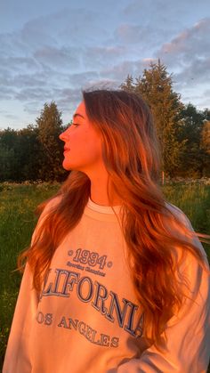 a girl with long red hair standing in the grass and looking off into the distance