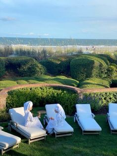 two women in white robes are sitting on lawn chairs near the beach and bushes with hedges