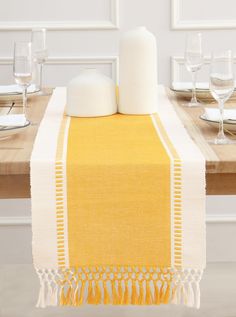 a yellow table runner with white tassels and two bottles on the top, next to wine glasses