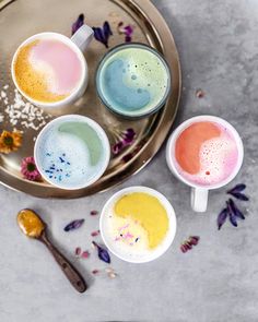 four different colored drinks on a metal tray with spoons and flowers around the cups