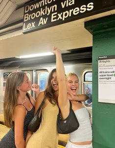 three women standing on a subway platform with their arms in the air