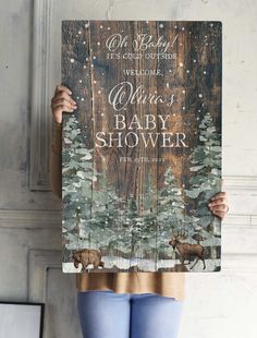 a woman holding up a wooden sign that says baby shower with deer and pine trees