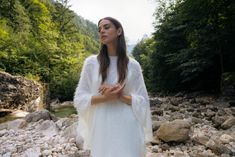 a woman standing in front of rocks and water wearing a white dress with long sleeves