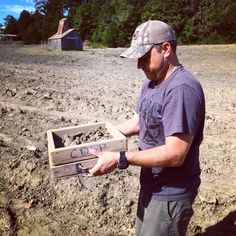 a man holding a wooden box in the middle of a dirt field with trees behind him