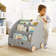 a toddler standing in front of a toy book rack with books on it and holding onto the top shelf