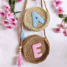 two straw bags with letters on them and tassels hanging from the handles next to pink flowers