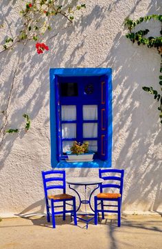 two chairs and a table in front of a white building with blue windows, one has a potted plant on the window sill