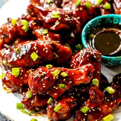 a white plate topped with wings covered in bbq sauce and green onions next to a small bowl of dipping sauce