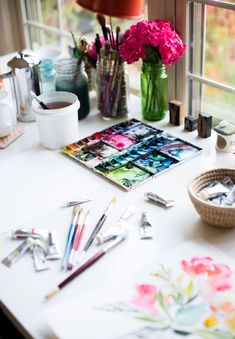 a table with art supplies and flowers in vases next to a window sill