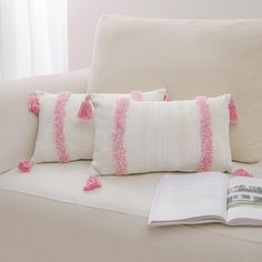 three pillows with pink tassels are sitting on a couch next to an open book