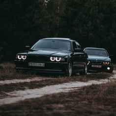 two black bmws parked on the side of a dirt road next to some trees