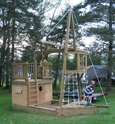 a child playing on a wooden pirate ship in the grass with trees behind him and another kid sitting at the top of it