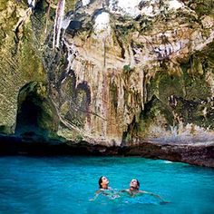 two people are swimming in the blue water near some rocks and cliffs, while another person is looking up at them