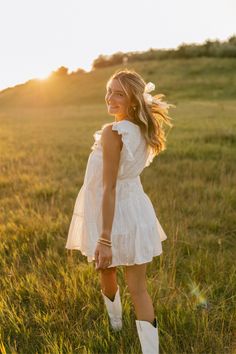 a woman in white dress and cowboy boots standing on grass with the sun behind her