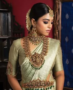 a woman in a green and gold sari with necklaces on her neck, standing next to a red wall