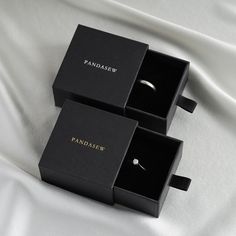 two black boxes sitting on top of a white bed covered in sheets and pillows, one with an engagement ring inside the box