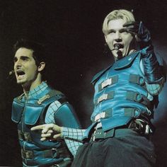 two men in blue costumes are performing on stage with one holding his hand up to the other