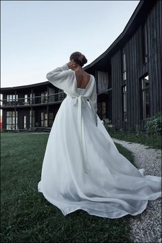 a woman in a long white dress is standing on the grass near a building and looking off into the distance