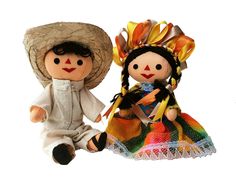 two dolls sitting next to each other on a white surface, one wearing a straw hat and the other in a colorful dress