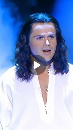 a man with long hair standing in front of a blue background and wearing a white shirt