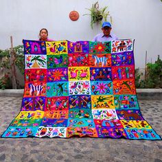 a man and woman holding up a brightly colored quilt on the ground in front of a white wall