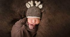 a newborn baby wearing a knitted deer hat and sleeping on a brown blanket with his hands under his chin