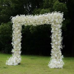 a white flower covered archway in the middle of a grassy area with trees behind it