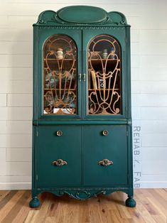 a green china cabinet with glass doors and gold trimmings on the top, in front of a white brick wall