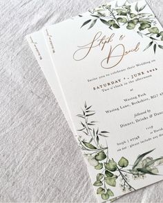 two wedding cards with greenery on them sitting on top of a white table cloth