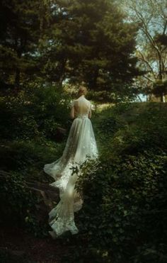 a woman in a wedding dress walking through the woods with her back to the camera