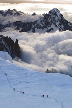 several skiers trekking up the side of a snow covered mountain with clouds in the background