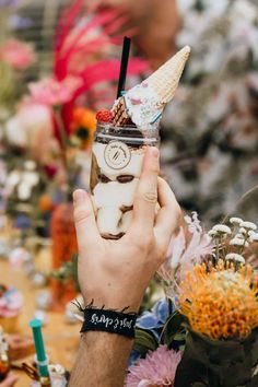 a person holding up a drink with an ice cream sundae on it and flowers in the background