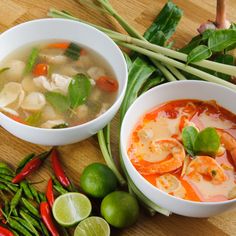 two white bowls filled with soup next to green vegetables and limes on a wooden table